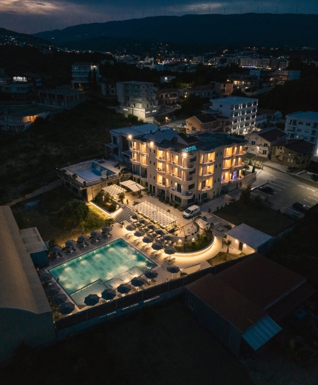Aerial view of Hotel Bluemoon Ulcinj at dusk, featuring an illuminated exterior and outdoor pool area with numerous sunbeds and umbrellas, surrounded by buildings and hills in the background.
